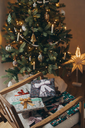 Merry Christmas! Stylish christmas gifts wrapped in festive paper with bows, vintage ornament and toy on modern chair on background of christmas tree lights. Atmospheric winter holidaysの写真素材