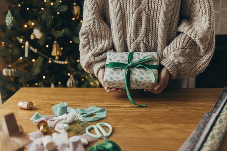 Woman in cozy sweater holding stylish christmas gift in festive wrapping paper with bow, ribbon, vintage ornaments on wooden table. Atmospheric winter holidays, wrapping christmas presentの写真素材