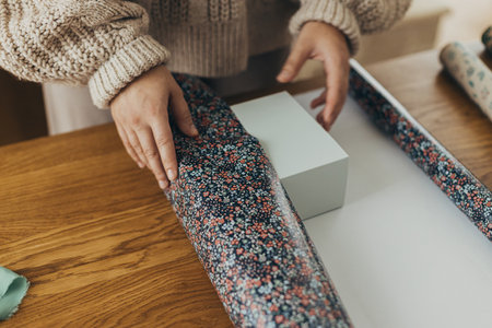 Woman in cozy sweater wrapping stylish christmas gift in festive wrapping paper with ribbons,...