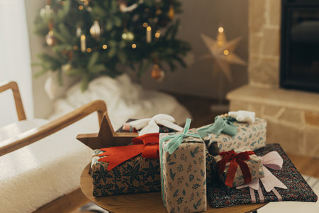 Merry Christmas! Stylish christmas gifts wrapped in festive paper with bows and vintage ribbons on wooden table on background of christmas tree lights. Atmospheric winter holidaysの写真素材