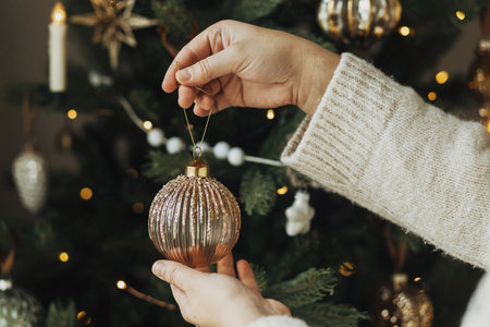 Decorating christmas tree with stylish bauble. Hands in cozy sweater hanging vintage ornament on tree branch close up. Atmospheric winter holiday tradition, family timeの写真素材