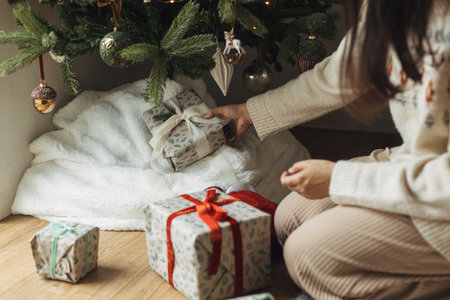 Woman in cozy sweater putting stylish christmas gifts under decorated christmas tree with modern vintage baubles. Atmospheric winter holidays time. Preparing Christmas presentsの写真素材