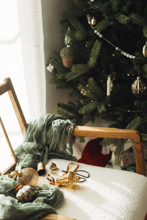 Stylish christmas vintage baubles, ribbons, brass bells and scissors on cozy white chair under tree. Christmas ornaments aesthetics. Decorating christmas tree. Merry Christmas!の写真素材