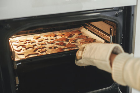 Woman in oven mitts putting tray with fresh gingerbread cookies in christmas festive shapes in modern oven in white kitchen. Making christmas gingerbread cookiesの写真素材