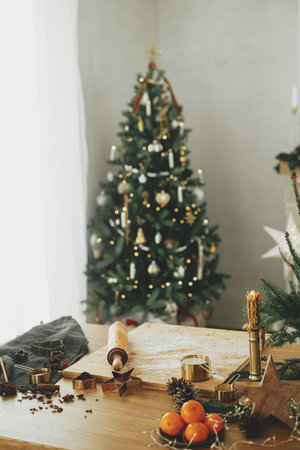 Flour on wooden board, rolling pin, golden metal cutters, cooking spices and festive decorations on rustic table against stylish decorated christmas tree. Making christmas gingerbread cookiesの写真素材
