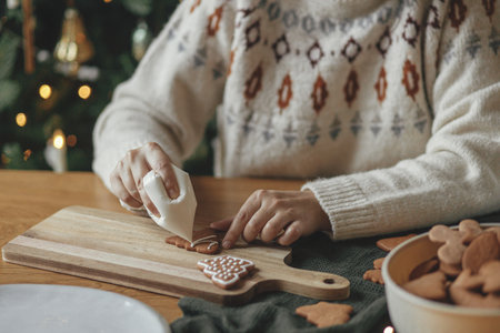 Decorating gingerbread cookies with icing on rustic wooden table close up. Atmospheric Christmas holiday traditions. Hands decorating baked christmas cookies with sugar frosting. Family timeの写真素材