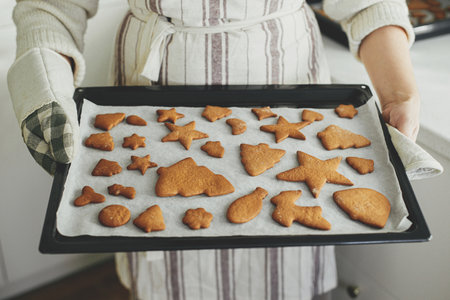 Woman in apron and oven mitts holding tray with fresh baked gingerbread cookies in christmas festive...