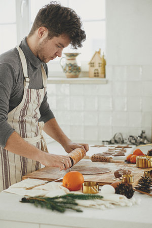 Man making christmas gingerbread cookies in modern white kitchen. Hand kneading gingerbread dough on wooden board with flour, rolling pin, golden metal cutters, cooking spices on countertopの写真素材