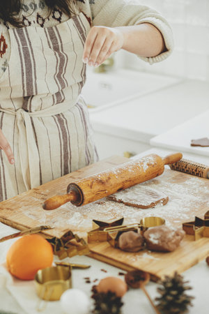 Hands kneading gingerbread dough on wooden board with rolling pin, golden metal cutters, cooking...