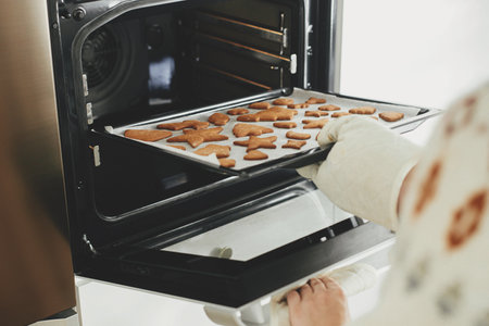Hands in oven mitts holding tray with fresh baked gingerbread cookies in christmas festive shapes in modern white kitchen. Making christmas gingerbread cookiesの写真素材