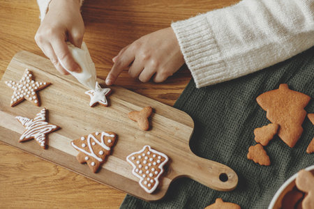 Decorating gingerbread cookies with icing on rustic wooden table top view. Atmospheric Christmas holiday traditions. Hands decorating baked christmas cookies with sugar frosting. Family timeの写真素材