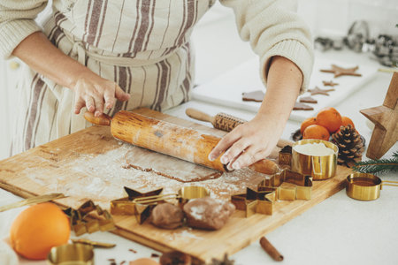 Hands kneading gingerbread dough on wooden board with rolling pin, golden metal cutters, cooking spices, festive decorations in modern white kitchen. Woman making christmas gingerbread cookiesの写真素材