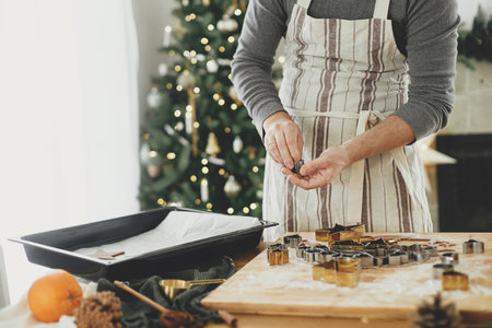 Hands holding raw gingerbread cookie in festive shapes at baking tray on rustic table with holiday decorations. Man making christmas gingerbread cookies, atmospheric holiday timeの写真素材