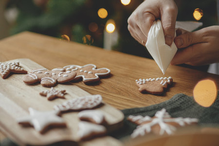 Decorating gingerbread cookies with icing on rustic wooden table at christmas tree golden lights. Atmospheric Christmas holiday traditions. Man decorating cookies with sugar frostingの写真素材