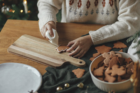 Decorating gingerbread cookies with icing on rustic wooden table close up. Atmospheric Christmas holiday traditions. Hands decorating baked christmas cookies with sugar frosting. Family timeの写真素材