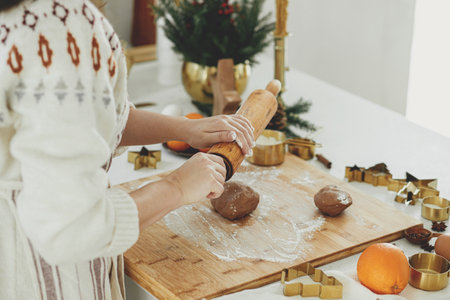 Hands kneading gingerbread dough on wooden board with rolling pin, golden metal cutters, cooking spices, festive decorations in modern white kitchen. Woman making christmas gingerbread cookiesの写真素材