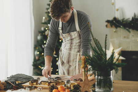 Hands cutting gingerbread dough with festive cutters on rustic table with holiday decorations against stylish christmas tree. Man making christmas gingerbread cookies, atmospheric holiday timeの写真素材