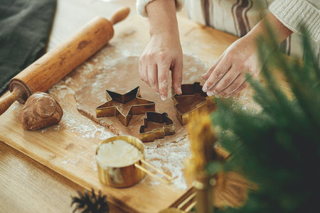 Hands cutting gingerbread dough with festive golden cutters on rustic table with holiday decorations, close up. Woman making christmas gingerbread cookies, atmospheric timeの写真素材