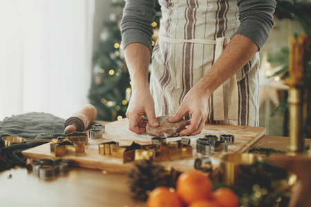 Hands kneading gingerbread dough with rolling pin, cooking spices, festive decorations on rustic table against stylish christmas tree. Man making christmas gingerbread cookies, holiday timeの写真素材