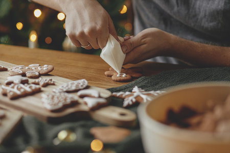 Decorating gingerbread cookies with icing on rustic wooden table at christmas tree golden lights. Atmospheric Christmas holiday traditions. Man decorating cookies with sugar frostingの写真素材