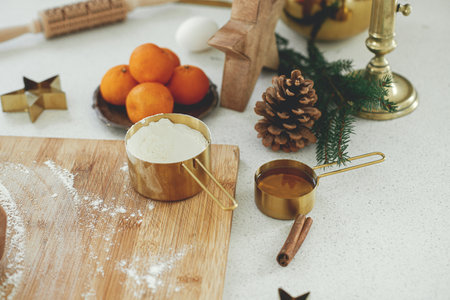 Making christmas gingerbread cookies in modern white kitchen. Gingerbread dough on wooden board, flour, rolling pin, golden metal cutters, cooking spices and festive decorations on countertopの写真素材