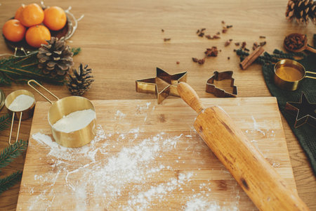 Making christmas gingerbread cookies. Flour on wooden board, rolling pin, golden metal cutters, cooking spices and festive decorations on rustic tableの写真素材