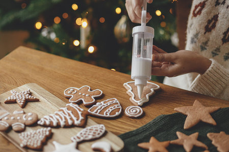 Decorating gingerbread cookies with icing on rustic wooden table at christmas tree golden lights. Atmospheric Christmas holiday traditions. Woman decorating cookies with sugar frostingの写真素材