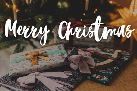 Merry Christmas text on stylish christmas wrapped gifts on background of christmas tree lights. Happy Holidays! Season's greeting card. Handwritten signの写真素材