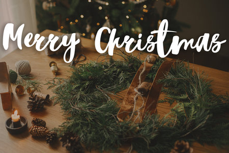 Merry Christmas text on stylish christmas rustic wreath with cedar branches, ribbon, vintage bells on table. Happy Holidays! Season's greeting card. Handwritten signの写真素材