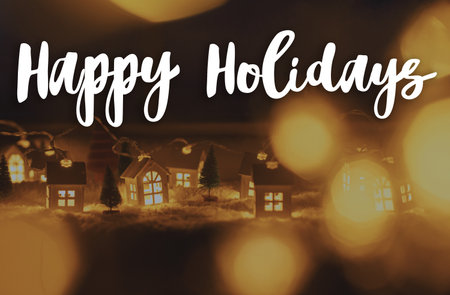 Happy Holidays text on cozy christmas miniature village with lights bokeh in evening. Merry Christmas. Season's greeting card. Handwritten signの写真素材