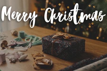 Merry Christmas text on stylish christmas gift, festive wrapping paper, ribbons, vintage ornaments against christmas tree. Happy Holidays! Season's greeting card. Handwritten signの写真素材