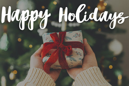 Happy Holidays text on hands in cozy sweater holding stylish christmas gift with red ribbon at christmas tree with lights. Merry Christmas. Season's greeting card. Handwritten signの写真素材