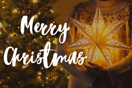 Merry Christmas text on stylish illuminated golden star in hands on background of christmas tree with golden lights in evening. Happy Holidays! Season's greeting card. Handwritten signの写真素材
