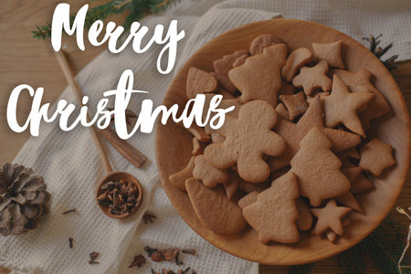 Merry Christmas text on christmas gingerbread cookies in wooden plate on table flat lay with fir branches, pinecone, spices. Happy Holidays! Season's greeting card. Handwritten signの写真素材