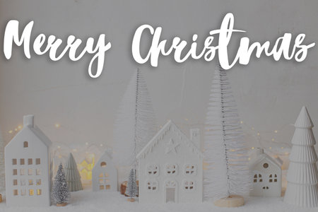 Merry Christmas text on stylish little white houses and trees, snowy christmas miniature village  with golden lights. Happy Holidays! Season's greeting card. Handwritten signの写真素材