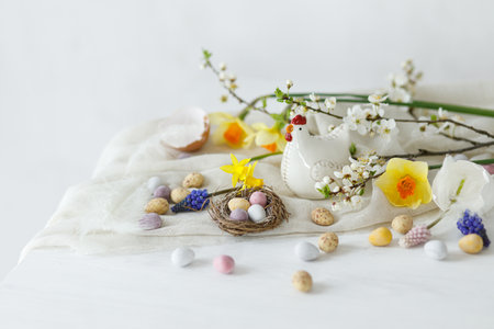 Happy Easter! Stylish easter chocolate eggs in nest, spring flowers, chicken figurine on white rustic wooden table. Easter modern simple banner, space for text. Seasons greetingsの写真素材