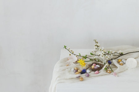 Happy Easter! Stylish easter chocolate eggs in nest, spring flowers, feathers and linen cloth on white rustic wooden table. Easter modern simple banner, space for text. Seasons greetingsの写真素材