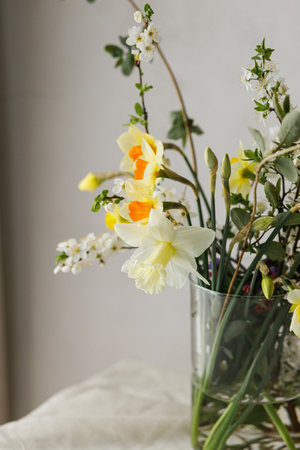 Stylish spring flowers bouquet on rustic table in rural room. Beautiful daffodils, cherry bloom and greenery composition in glass vase. Easter modern simple decorの写真素材