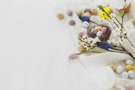Happy Easter! Stylish easter chocolate eggs in nest, spring flowers, feathers and linen cloth on white rustic wooden table. Easter modern simple banner, space for text. Seasons greetingsの写真素材
