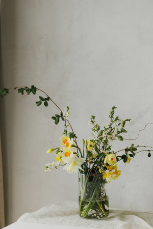 Beautiful daffodils, cherry bloom and greenery composition in glass vase. Stylish spring flowers bouquet on rustic table in rural room. Easter modern simple decorの写真素材