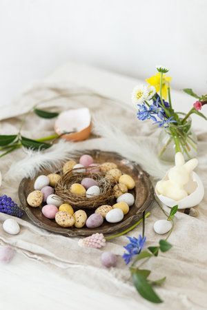 Stylish easter chocolate eggs in nest, spring flowers, feathers and linen cloth on rustic wooden table. Easter modern simple decoration still life. Happy Easter!の写真素材