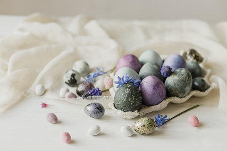 Happy Easter! Stylish easter eggs in modern tray and spring flowers on linen cloth on rustic white table. Easter simple banner, space for text. Natural painted marble and purple eggs still lifeの写真素材