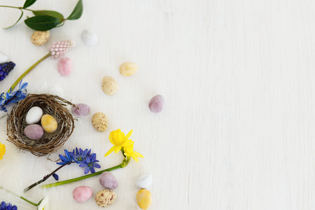 Easter rustic flat lay. Colorful easter chocolate eggs in nest, spring flowers, feathers border composition on white wooden table. Space for text. Happy Easter! Seasons greetingsの写真素材