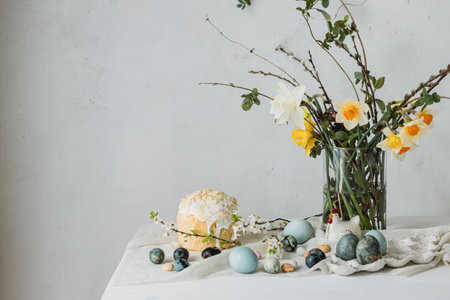 Happy Easter! Stylish easter eggs, homemade easter bread and spring flowers on linen napkin on rustic table against wall. Natural painted blue and marble eggs and daffodils bouquet still lifeの写真素材