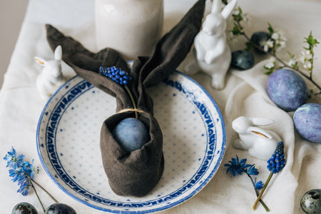 Happy Easter! Stylish easter egg in bunny napkin on vintage plate and spring flowers on linen napkin, rustic table setting. Natural painted blue eggs and hyacinth blooms. Modern minimal still lifeの写真素材