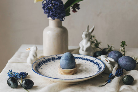 Stylish easter table setting. Easter egg in bunny figurine on vintage plate and spring flowers on rustic table. Happy Easter! Natural dye blue eggs and purple hiacynt. Holiday minimal still lifeの写真素材