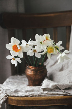 Stylish daffodils in vintage vase on old wooden chair with linen cloth.  Rustic still life. Countryside moody spring flowers composition. Easter floral bannerの写真素材