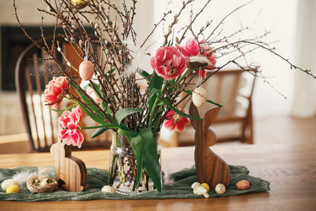 Stylish tulips bouquet with willow branches, easter eggs and wooden bunny figurines decor on wooden table close up. Happy Easter! Simple easter decoration in farmhouse living roomの写真素材