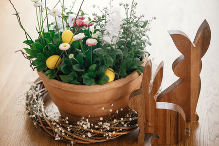 Stylish easter eggs with feather, potted spring flowers and bunny figurines composition on wooden table. Easter hunt. Simple easter table setting decor in farmhouse living roomの写真素材