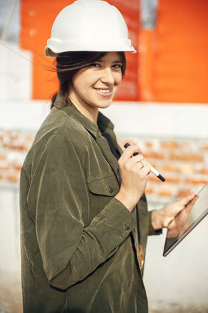 Happy young female architect with tablet checking blueprints against of building new modern house. Stylish happy woman engineer in hard hat looking at digital plans at construction siteの写真素材